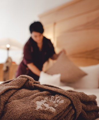 Dunkler Rehlegg-Bademantel liegt ordentlich gefaltet auf einem Bett während eine Mitarbeiterin die Kissen des Bettes richtet