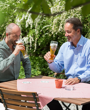 Zwei glückliche Männer sitzen bei strahlendem Sonnenschein im Biergarten und genießen ihr kühles Bier