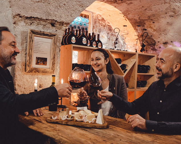 Drei Gäste stoßen im Weinkeller ihre Rotweingläser glücklich aneinander, vor Ihnen steht eine Käseplatte