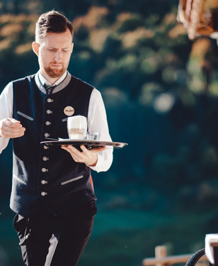 Kellner in Tracht serviert bei Sonnenschein einen Latte Macchiato