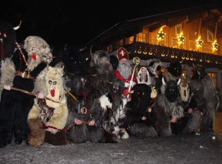 Zu Nikolaus verkleidete Krampusse posieren für ein Foto vor einer weihnachtlich beleuchteten Hütte